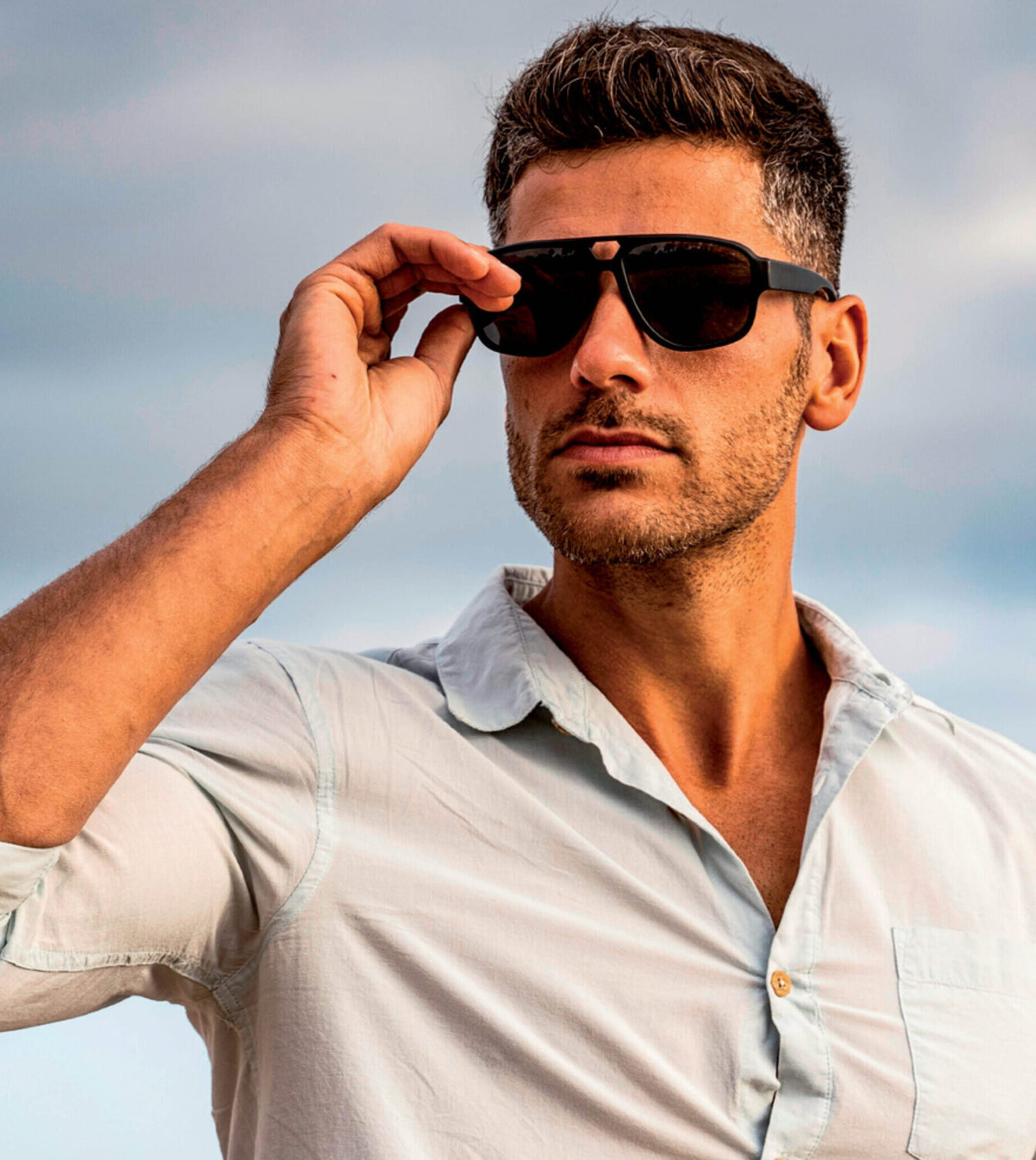 Een man met een stoppelbaard draagt stijlvolle zwarte zonnebrillen en een lichtblauw overhemd in de buitenlucht.