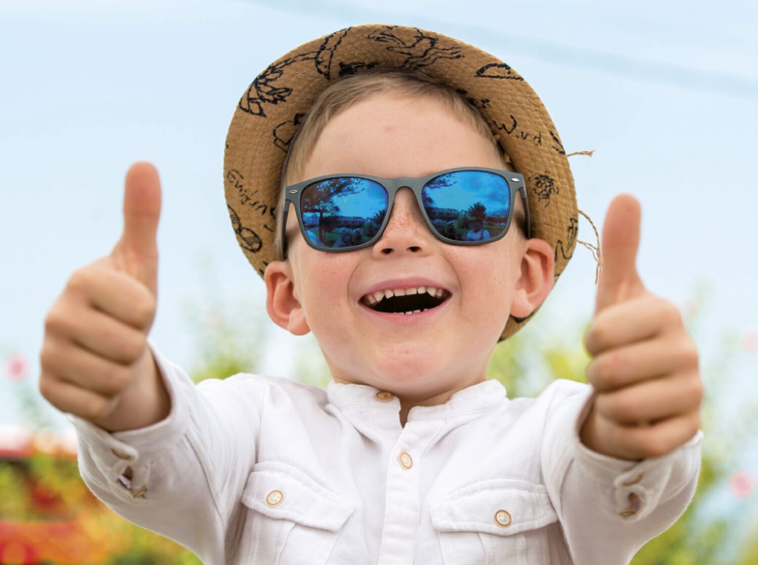 ein Junge mit rosigen Wangen, Strohhut und blauer Kindersonnenbrille zeigt den Daumen nach oben