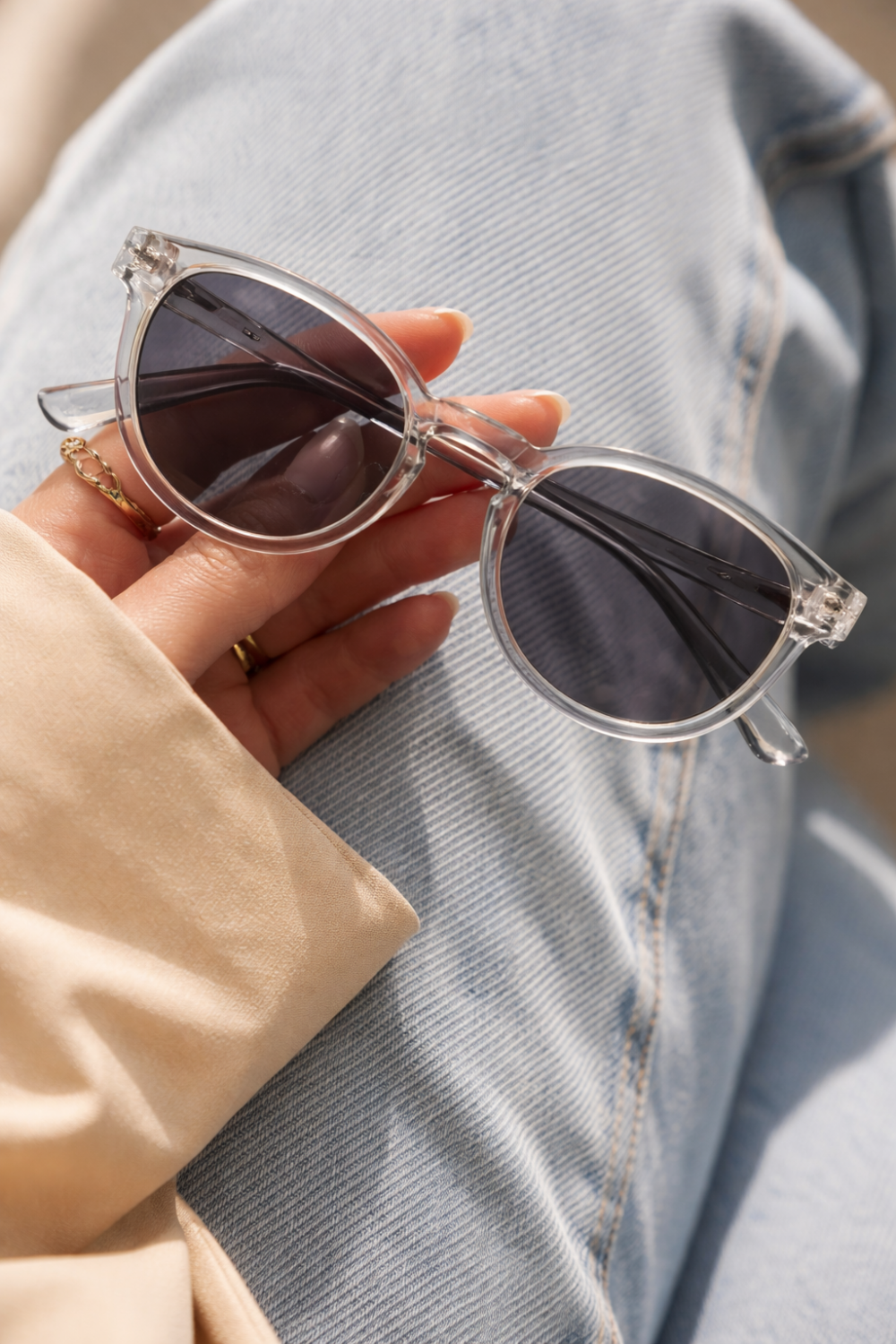 Eine Hand hält eine stylische Sonnenbrille aus transparentem Polycarbonat vor einem Hintergrund aus Jeans.