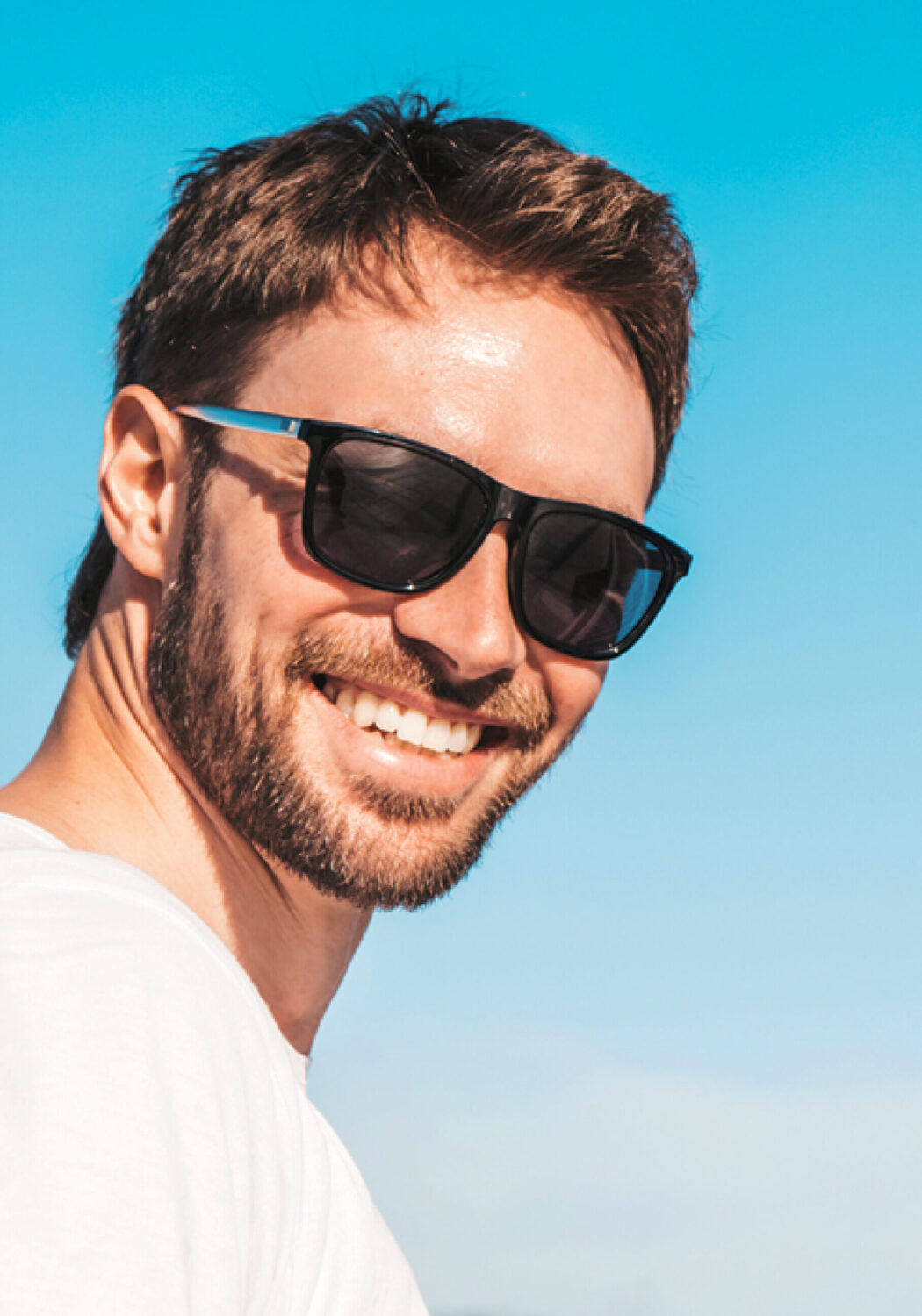 Lächelnder Mann mit Revex-Sonnenbrille, weißem T-Shirt und offenem Jeanshemd vor grauem Hintergrund