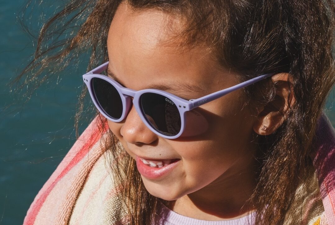 Lächelndes Mädchen mit lila Kindersonnenbrille von Okky und Strandtuch am Wasser, ideal für den Großhandel.