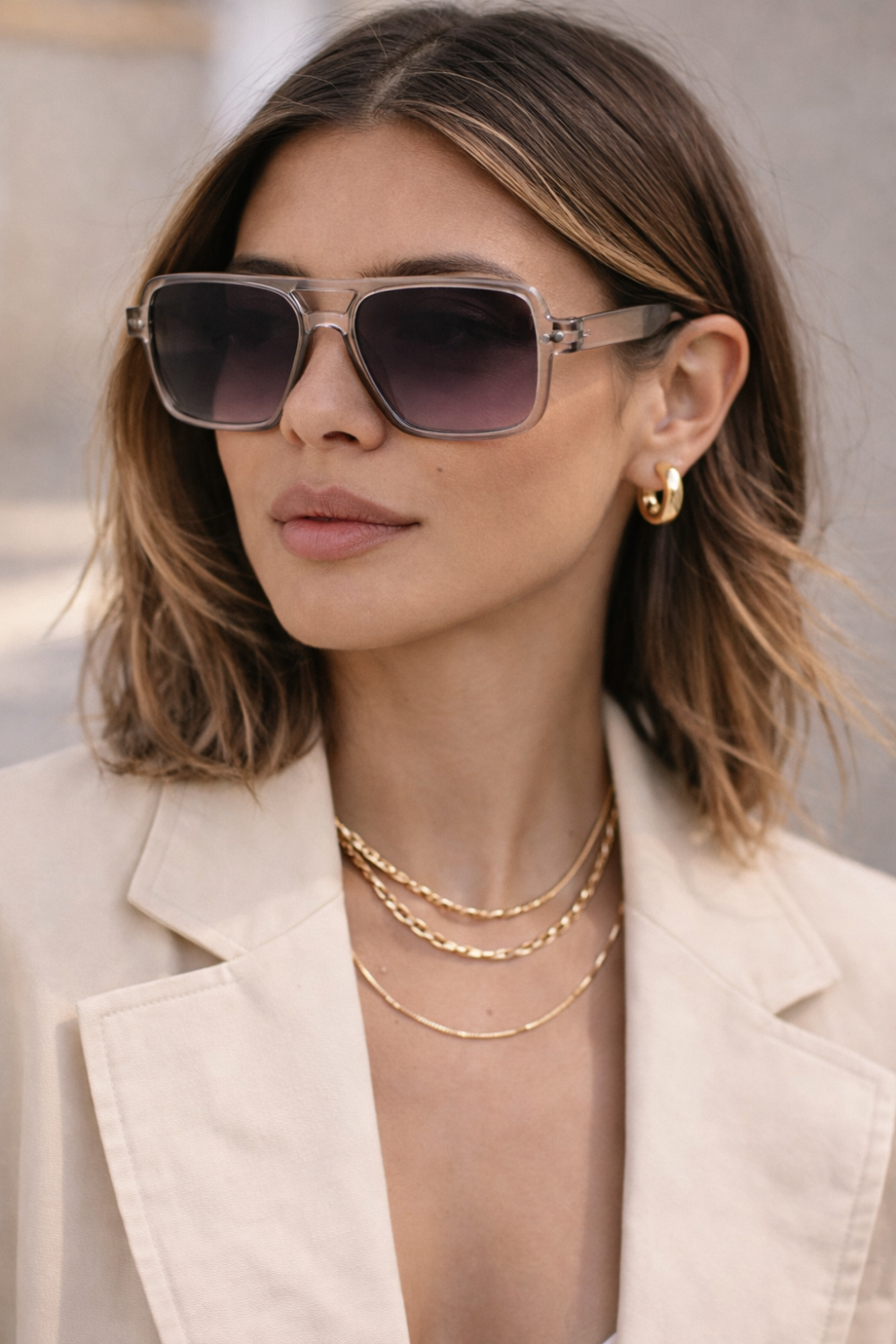 Femme élégante portant des lunettes en polycarbonate transparent, un blazer beige et des bijoux en or.