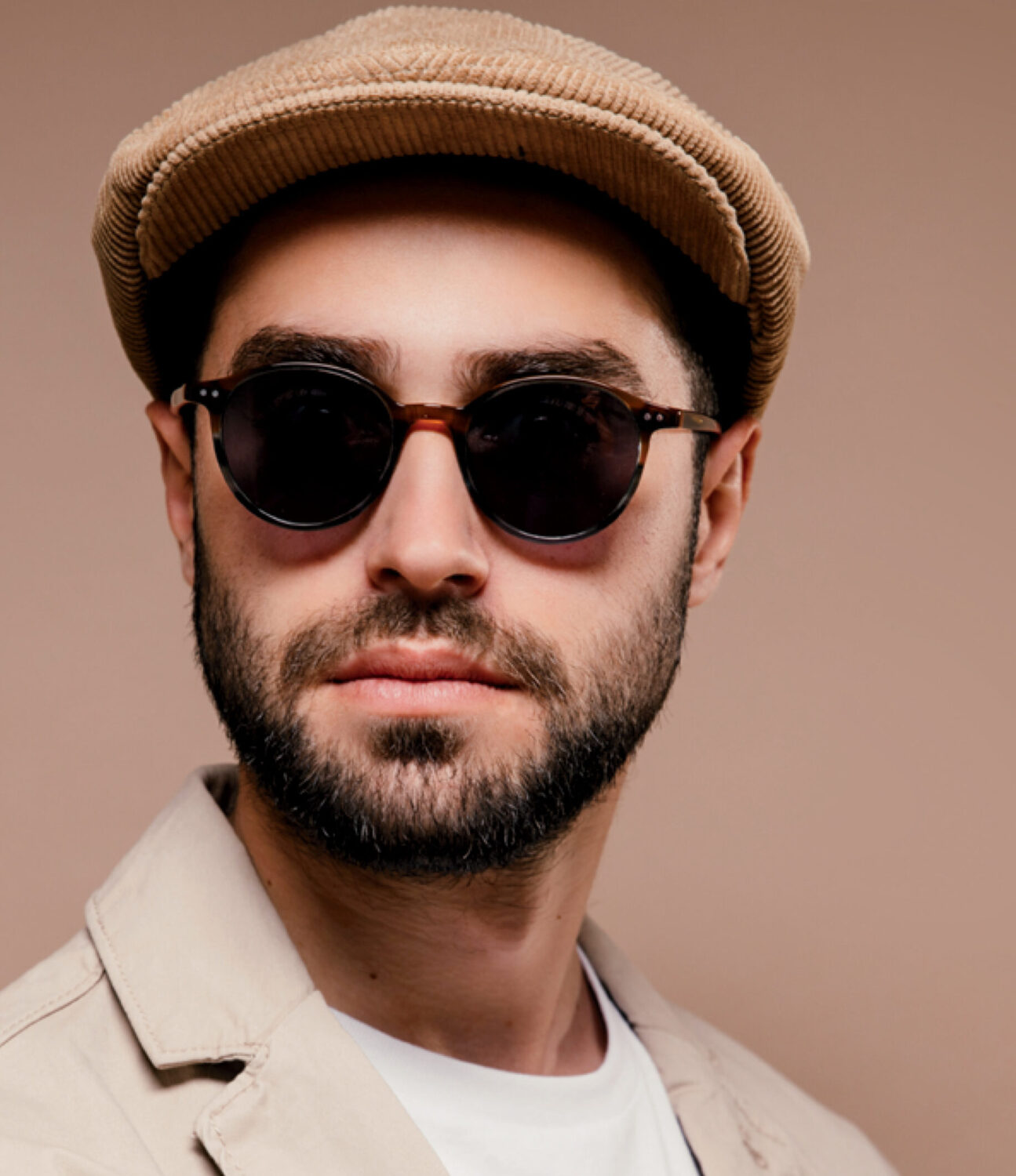 Homme barbu portant des lunettes de soleil rondes Level One et une casquette en velours côtelé beige sur un fond uni.