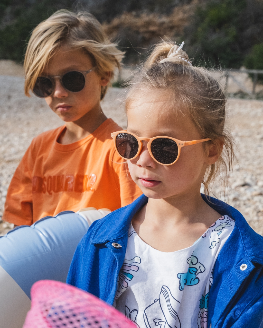 Un garçon et une fille blonds portent des lunettes de soleil pour enfants tendance de la marque Okky sur une plage de galets lors d'une journée ensoleillée.