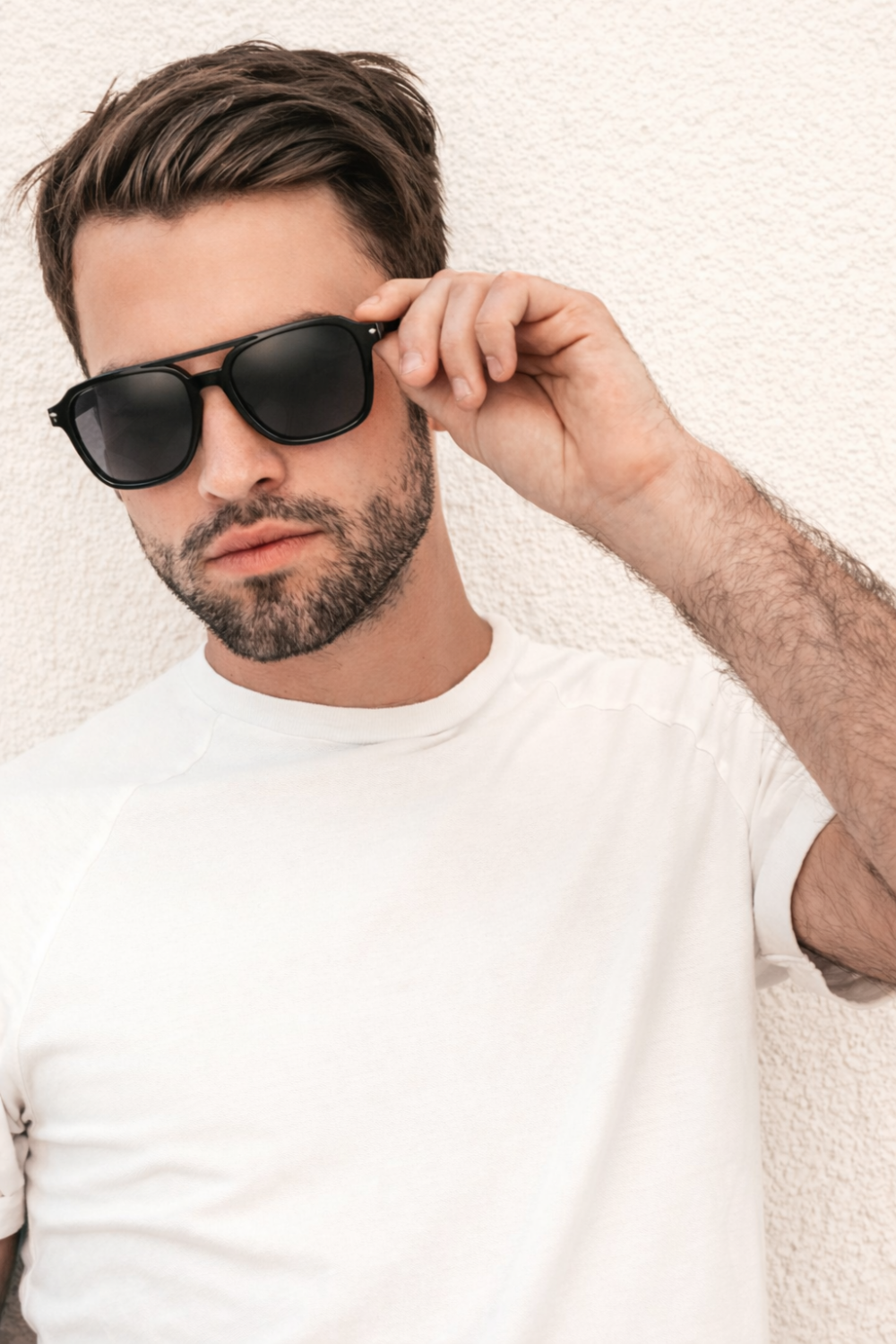 Homme stylé portant des lunettes de soleil noires et un t-shirt blanc contre un mur, focus sur les lunettes de soleil de l'homme.