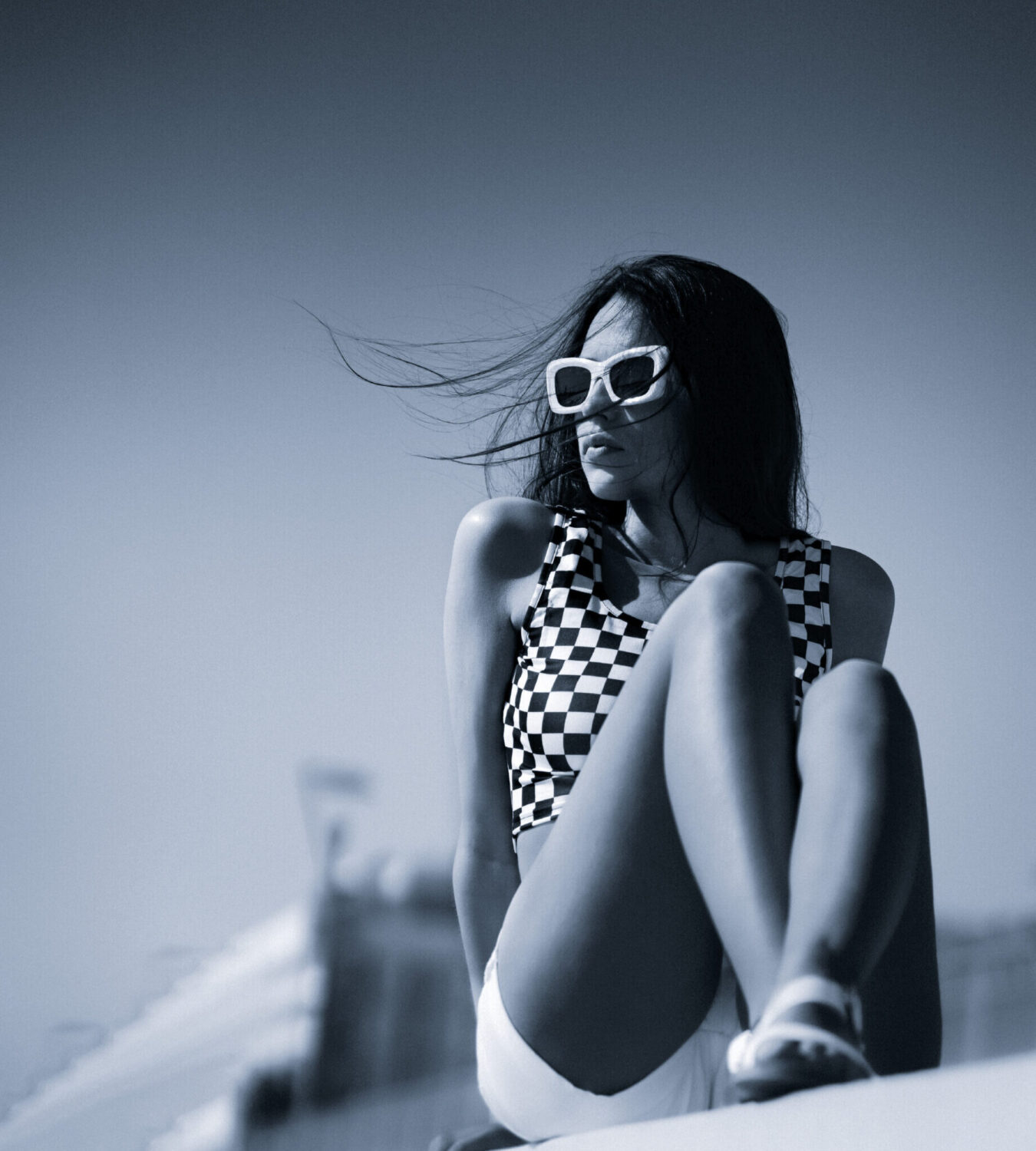 Mujer con gafas de sol blancas y top a cuadros sentada al aire libre con el pelo al viento.