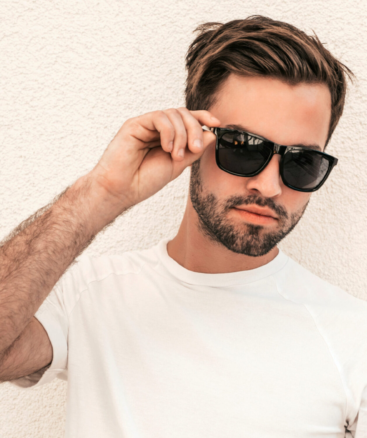 Hombre con gafas de sol negras Revex y camiseta blanca contra una pared blanca con textura.