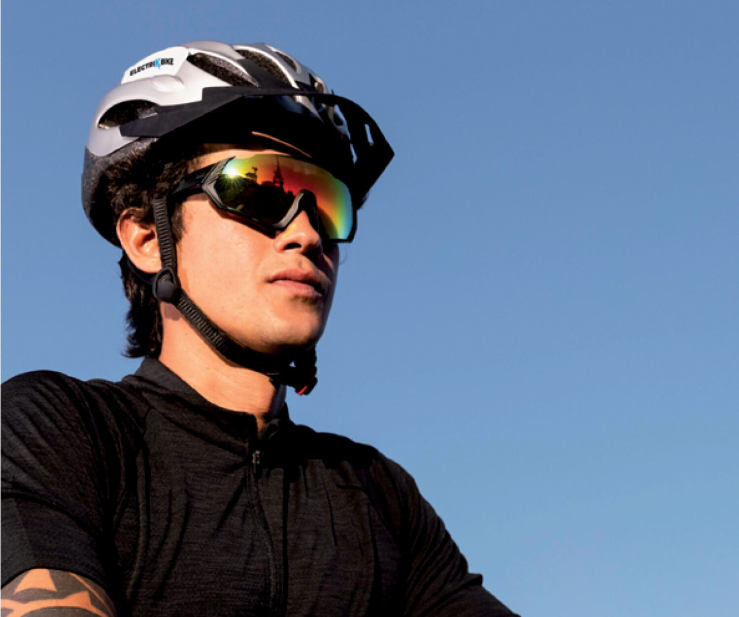 Hombre en bici con gafas de sol Revex Sport, casco de ciclista y tatuajes en los brazos contra un cielo azul despejado.