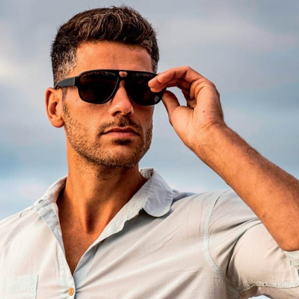 Un hombre elegante con camisa azul claro se endereza las gafas de sol negras contra un cielo despejado.