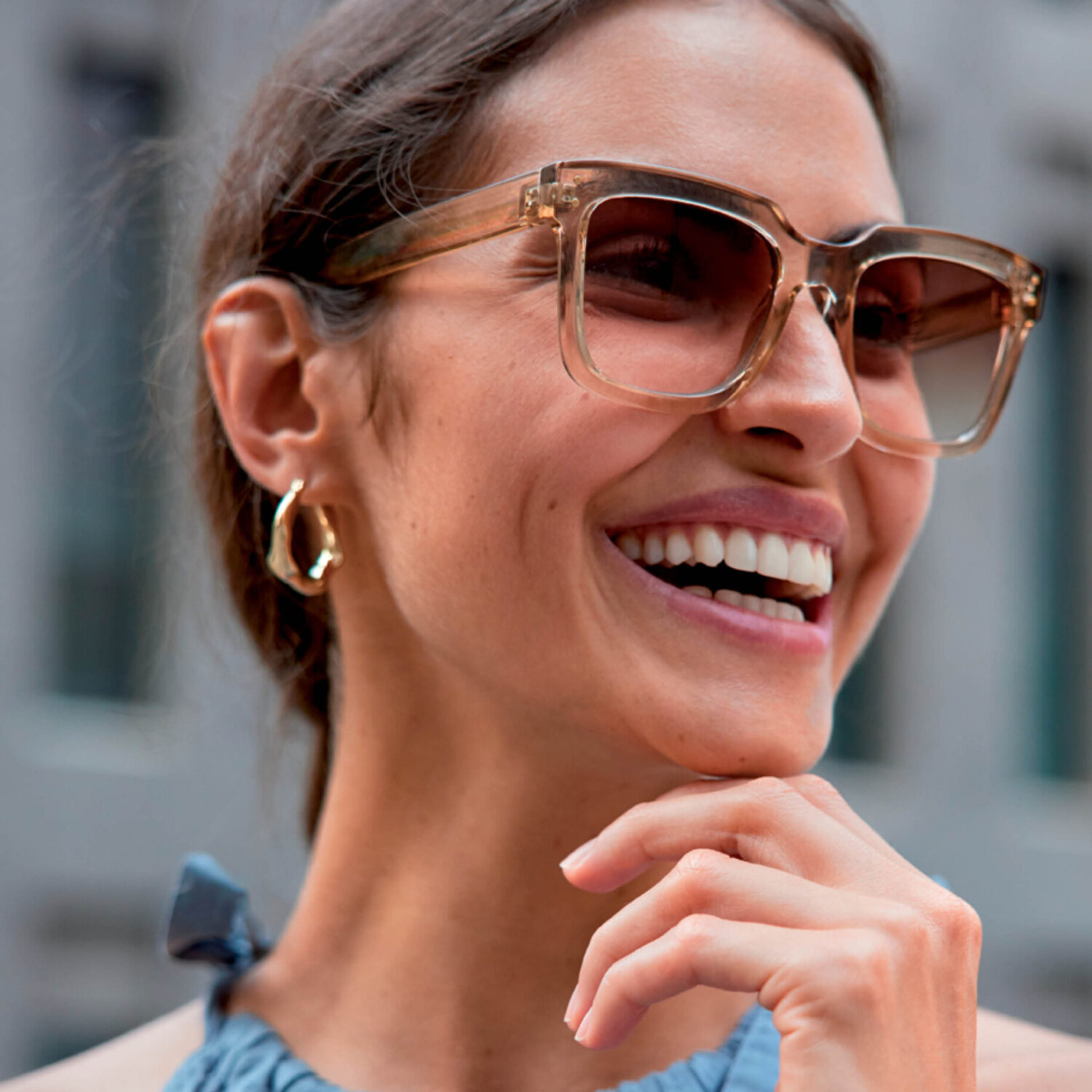 Smiling young woman wearing fashionable transparent brown sunglasses and gold earrings in an urban setting.