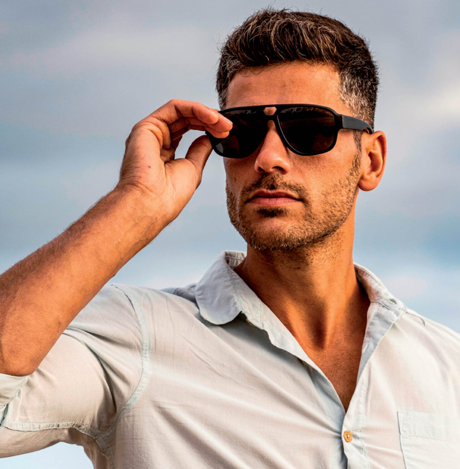 Man wearing stylish black Revex sunglasses and a light blue shirt outside against an overcast sky.
