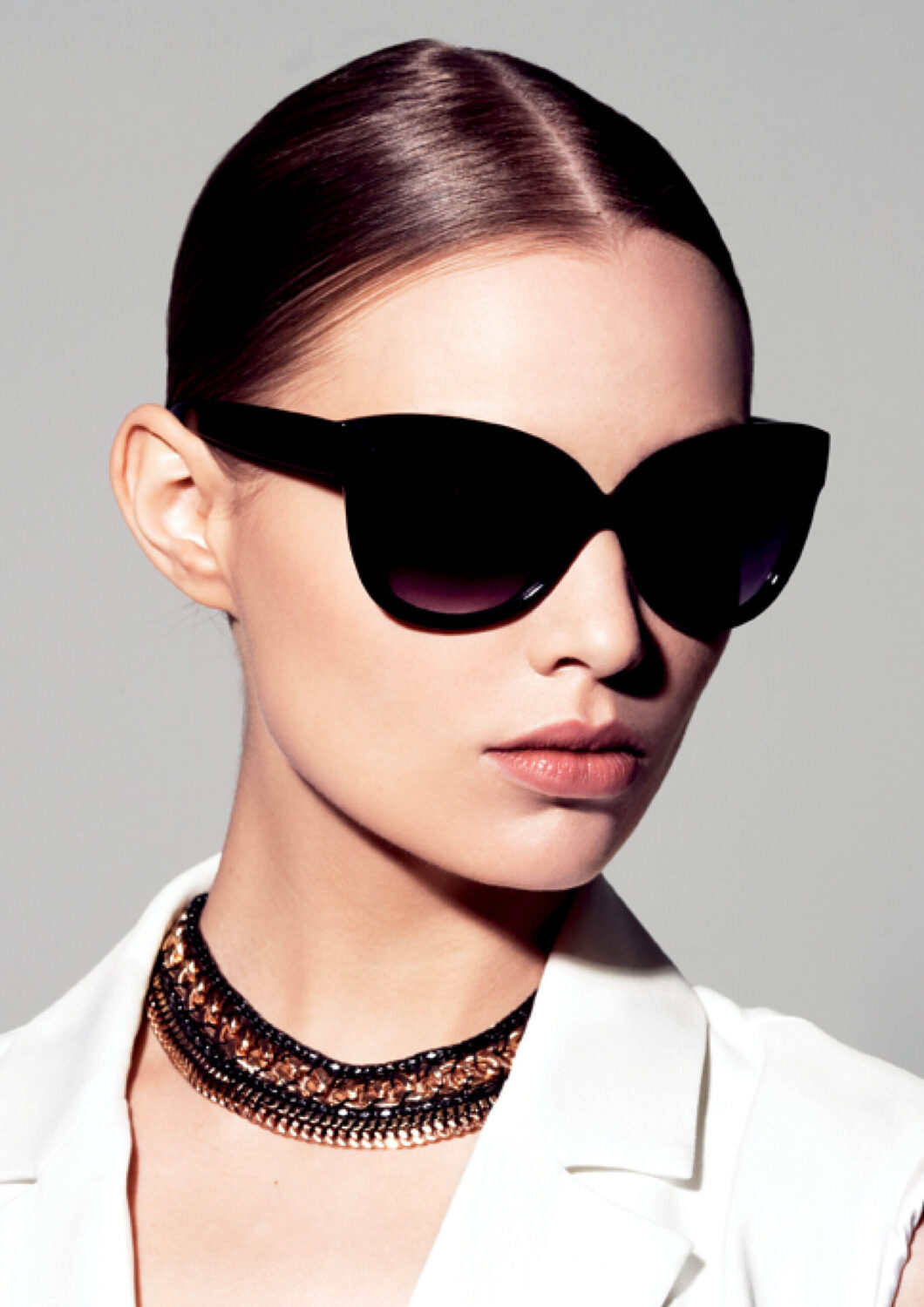 Stylish woman wearing large black Revex sunglasses and a gold-tone link chain against a grey background.