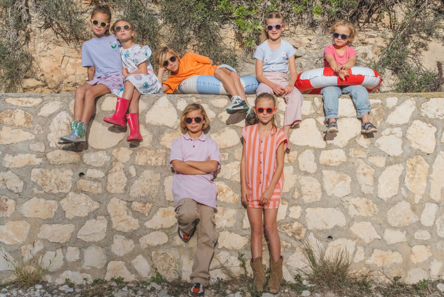 Group of happy children with sunglasses OKKY sitting and standing by a stone wall in the sun.