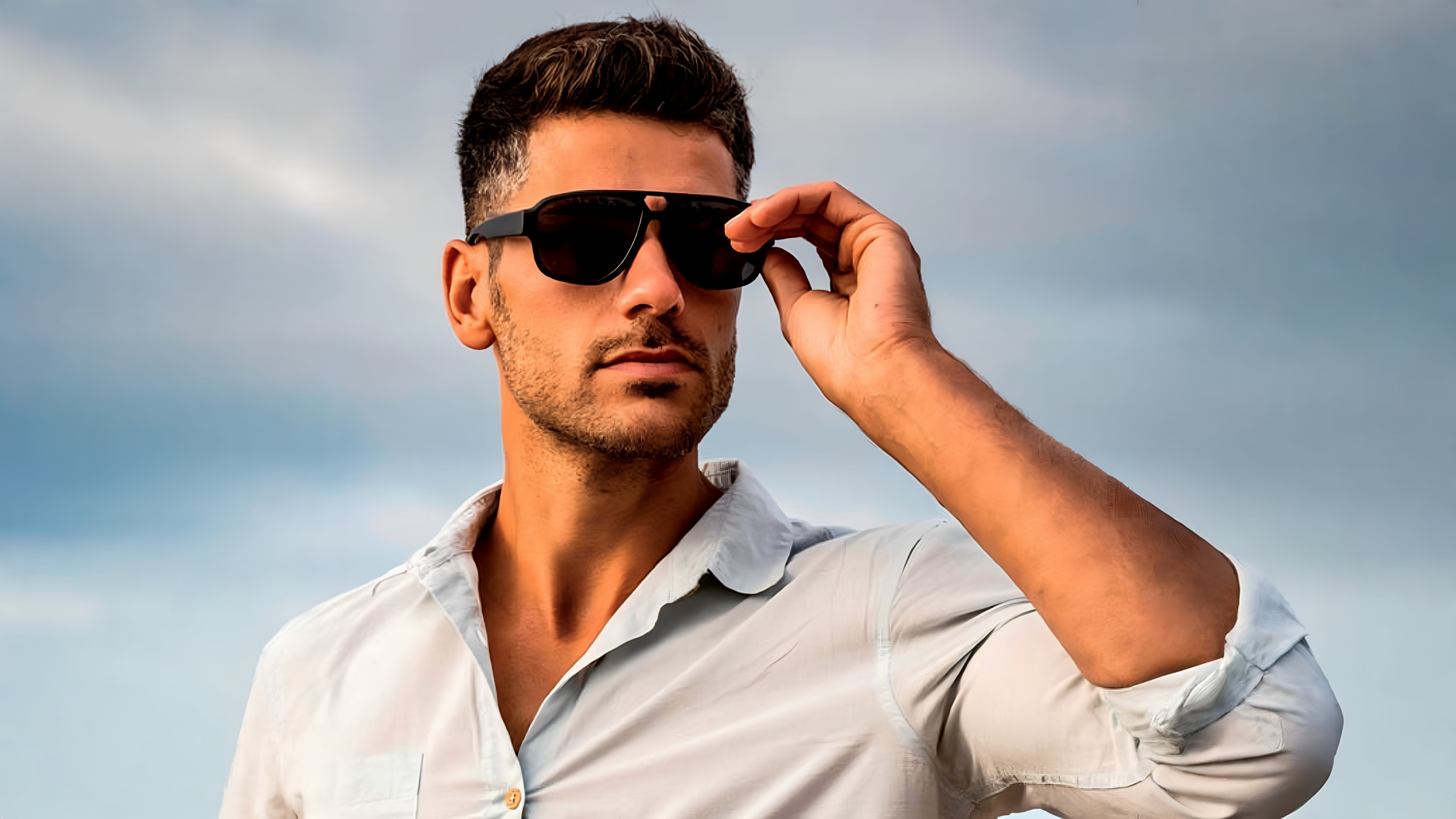 A man with stubble matches his black sunglasses against a cloudy blue sky.