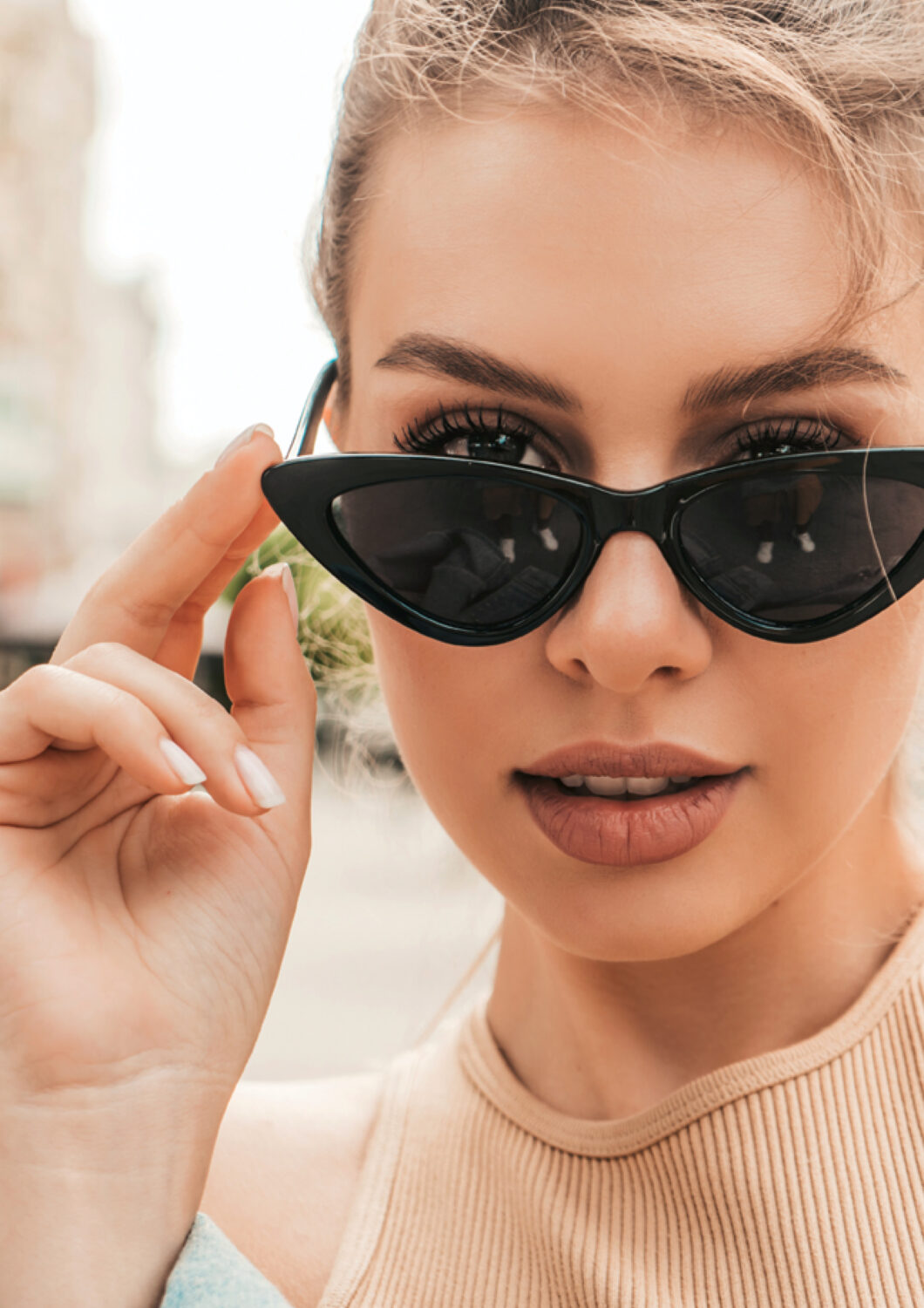 Blonde woman wearing black Revex sunglasses in cat-eye model looks over the frames in an urban setting.